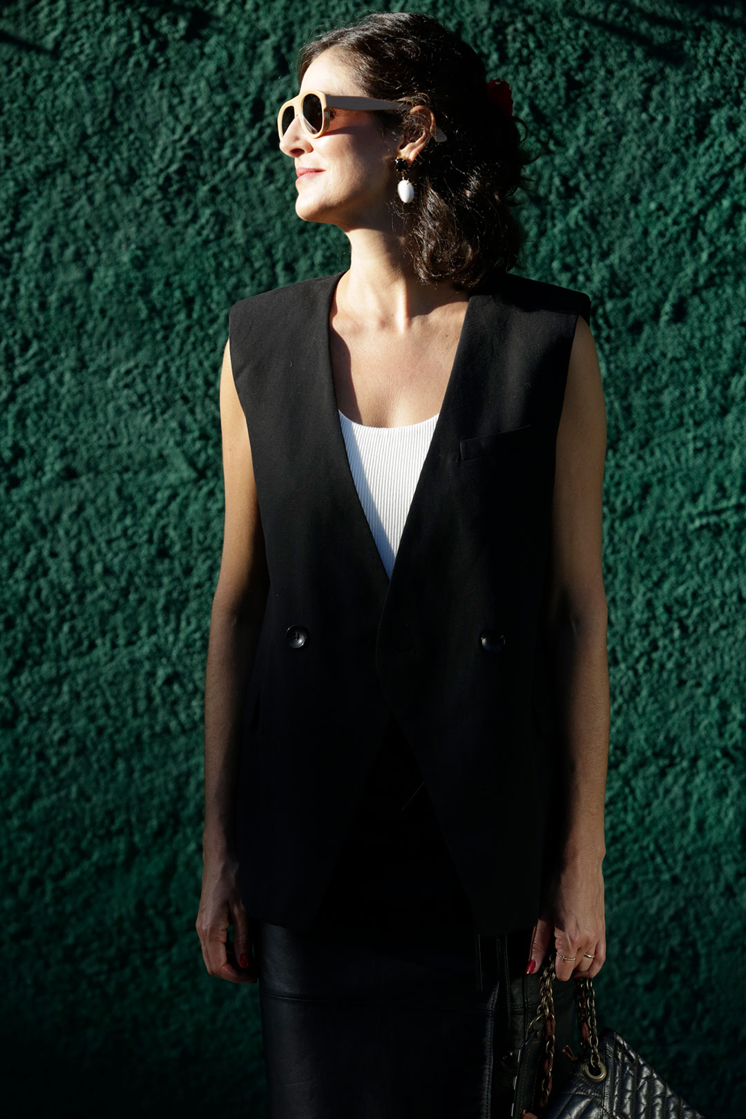 Luciana Chehin. Mulher sorridente de perfil usando óculos escuros de armação bege, colete preto longo sobre blusa branca e saia de couro preta. Ela segura uma bolsa preta acolchoada com alça de corrente em frente a uma parede verde texturizada sob luz solar forte