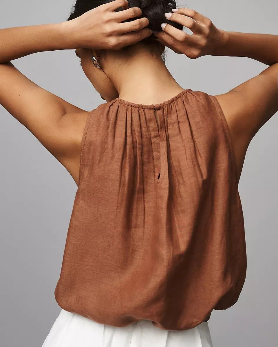 Vista posterior de uma pessoa usando uma blusa sem mangas na cor marrom terracota, com gola alta franzida e detalhe de fechamento por botão na nuca. O tecido possui textura de linho e a peça está combinada com uma parte inferior branca.