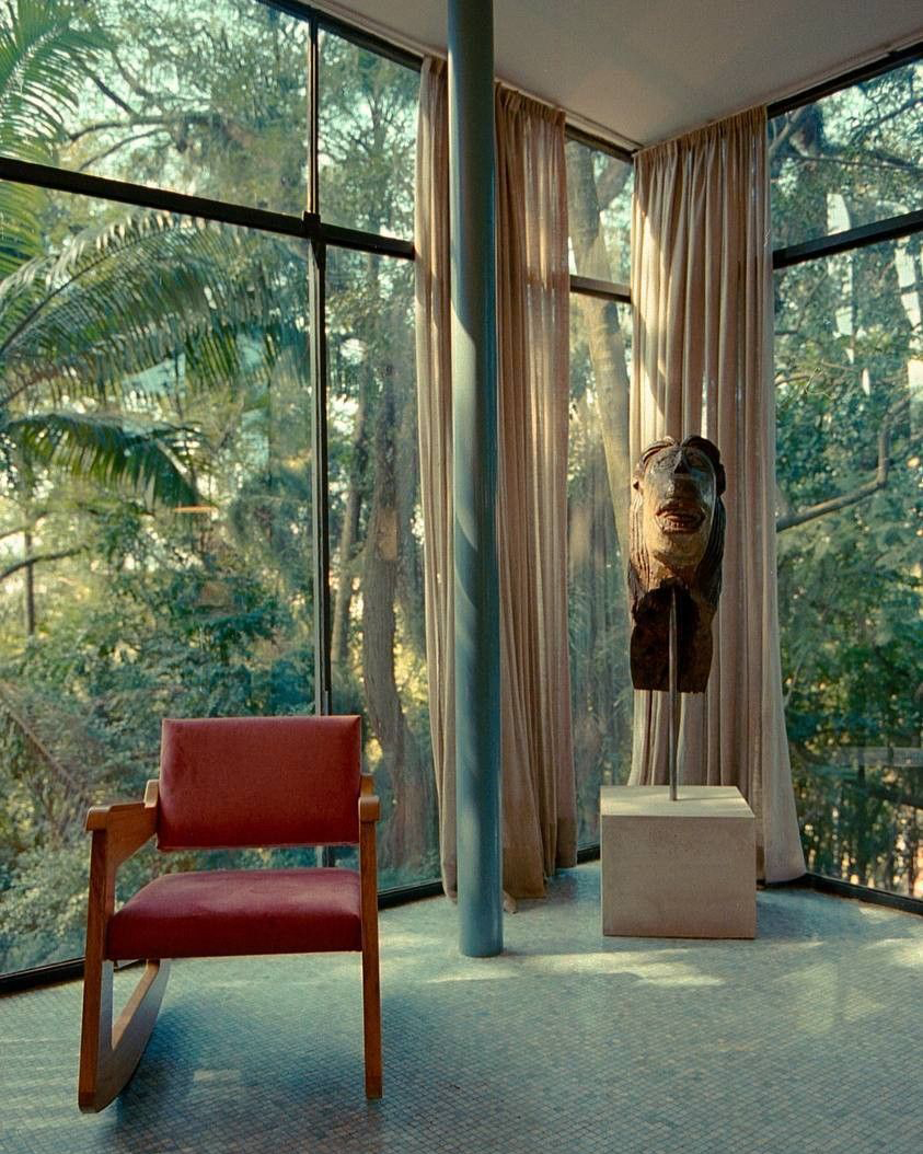 "Interior da Casa de Vidro de Lina Bo Bardi, com grandes fachadas de vidro que integram o ambiente à natureza externa. Em destaque, uma poltrona de balanço vermelha, uma coluna cilíndrica azul e uma escultura de máscara sobre um pedestal, com luz natural filtrada por cortinas leves.